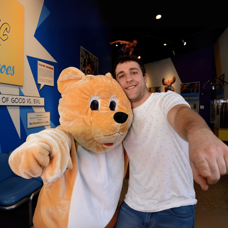 Person in a teddy bear costume posing with another man in front of Comic Book Heroes.