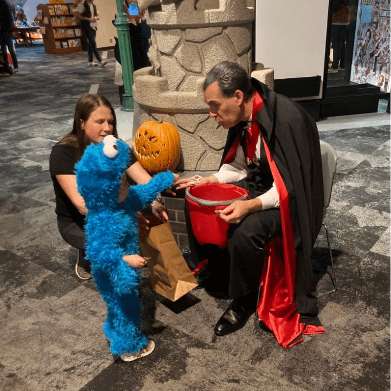 Child in a Cookie Monster costume reaching for a treat from a man in a "The Count" costume