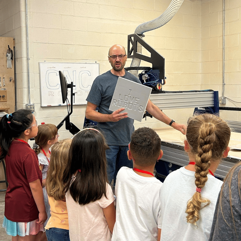 Campers standing in front of a man holding up a sign that reads "I Love Robots" as they learn about technology used by the exhibits team